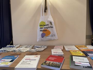 Büchertisch mit darüberhängender Tasche mit dem Leibniz-Zitat "die beste der möglichen Welten"